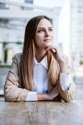 Thoughtful businesswoman with hand on chin sitting at cafe table - EBBF04720