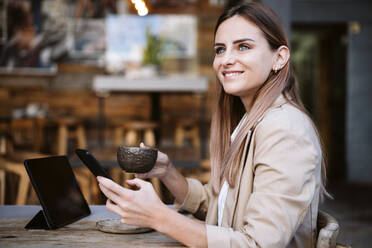 Lächelnde Geschäftsfrau mit Kaffeetasse und Smartphone auf der Terrasse eines Cafés - EBBF04724