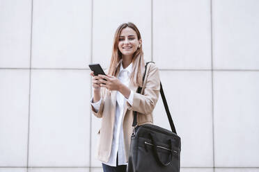 Lächelnde Geschäftsfrau mit Mobiltelefon und Umhängetasche vor einer weißen Wand - EBBF04737