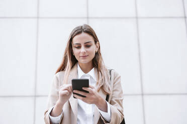 Female business professional using smart phone in front of wall - EBBF04739