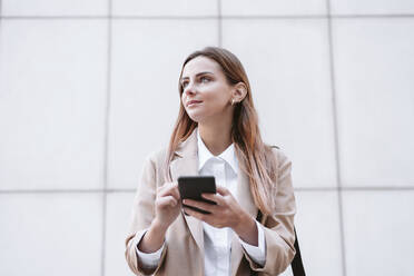 Businesswoman holding smart phone in front of wall - EBBF04740