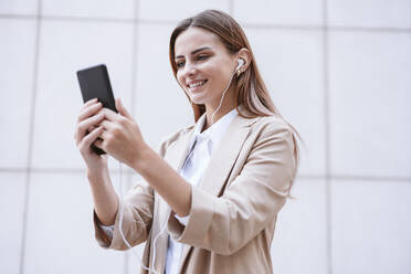 Businesswoman smiling while doing video call through smart phone in front of wall - EBBF04746