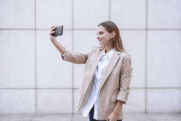 Young businesswoman taking selfie through smart phone in front of wall - EBBF04748