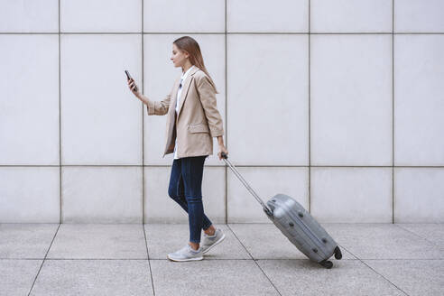 Geschäftsfrau, die ein Mobiltelefon benutzt, während sie mit Gepäck auf dem Fußweg geht - EBBF04750