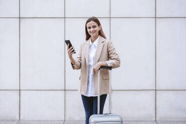 Smiling businesswoman with mobile phone and luggage in front of white wall - EBBF04751