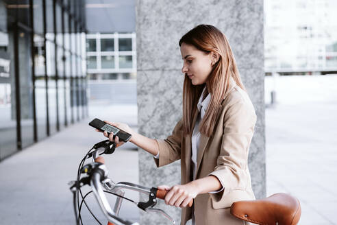 Geschäftsfrau entriegelt Elektrofahrrad per Smartphone an einer Parkstation - EBBF04759