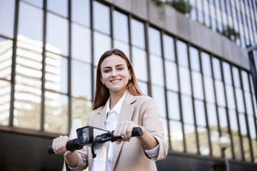 Smiling businesswoman with electric push scooter in city - EBBF04768