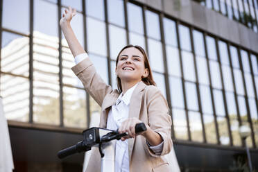 Businesswoman with electric push scooter raising hand in city - EBBF04769