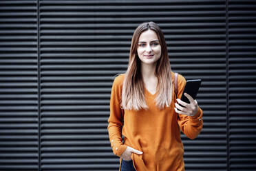 Young woman holding smart phone in front of wall - EBBF04773