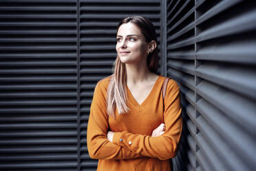 Young woman standing with arms crossed leaning on wall - EBBF04775