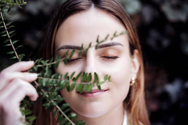 Lächelnde Geschäftsfrau mit geschlossenen Augen, die eine Pflanze vor das Gesicht hält - EBBF04797