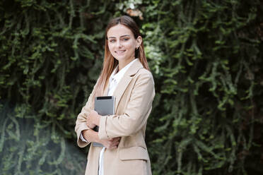 Beautiful smiling businesswoman standing with digital tablet by plants - EBBF04801