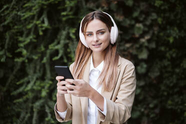 Geschäftsfrau, die ihr Smartphone in der Hand hält und über drahtlose Kopfhörer Musik hört - EBBF04803