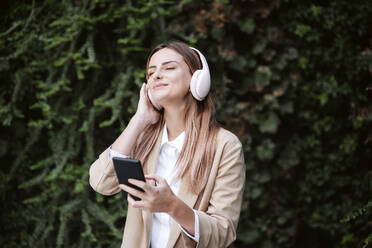 Weibliche Fachkraft mit geschlossenen Augen hört Musik über drahtlose Kopfhörer bei Pflanzen - EBBF04804