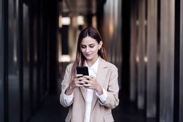 Geschäftsfrau, die im Flur stehend eine Textnachricht über ihr Mobiltelefon sendet - EBBF04811