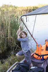 A boy hanging on to the canopy of a small boat on the water of the Okavango Delta - MINF16458