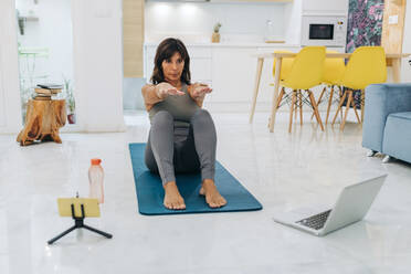 Mature female influencer doing yoga while vlogging in living room at home - JRVF01881