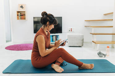 Reife Frau, die mit ihrem Smartphone Textnachrichten verschickt, während sie auf einer Übungsmatte im Wohnzimmer sitzt - JRVF01890
