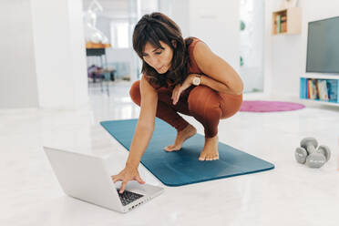 Woman crouching while using laptop on exercise mat - JRVF01894
