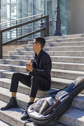 Junger männlicher Musiker spielt Gitarre, während er an einem sonnigen Tag auf einer Treppe sitzt - IFRF01064