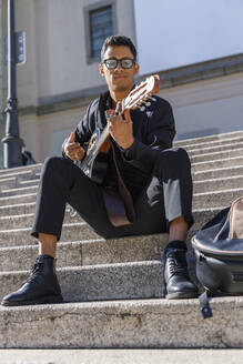 Männlicher Musiker mit Brille spielt Gitarre, während er auf einer Treppe sitzt - IFRF01066