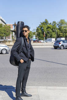 Junger Musiker, der einen Gitarrenkoffer trägt, während er mit den Händen in den Taschen auf der Straße an einem sonnigen Tag steht - IFRF01085