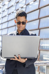 Junger Geschäftsmann mit Sonnenbrille, der einen Laptop benutzt, während er an einem sonnigen Tag am Geländer steht - IFRF01087