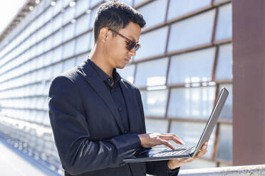 Männlicher Profi tippt auf einem Laptop, während er vor einem Bürogebäude steht - IFRF01088