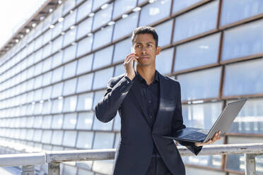 Young male professional with laptop talking on mobile phone while standing by railing - IFRF01090