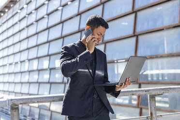 Geschäftsmann, der auf einen Laptop schaut, während er an einem sonnigen Tag am Geländer mit einem Smartphone spricht - IFRF01091