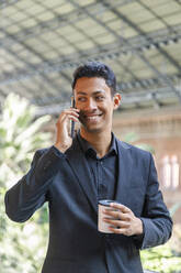 Glücklicher junger Geschäftsmann, der einen Einweg-Kaffeebecher hält und mit einem Mobiltelefon spricht - IFRF01097
