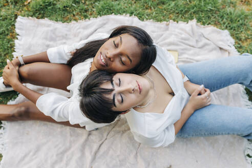 Freundinnen mit geschlossenen Augen sitzen Rücken an Rücken auf einer Decke - JRVF01917