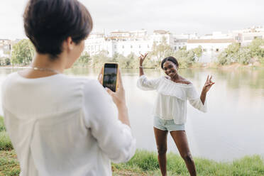 Woman photographing girlfriend gesturing at lakeshore - JRVF01930