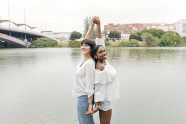 Lächelnde Frauen mit erhobenen Armen, die sich am See an den Händen halten - JRVF01937