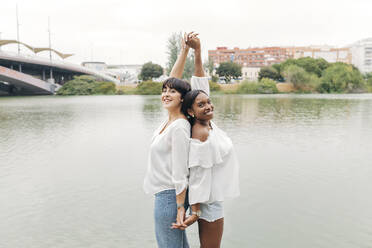 Smiling women with arms raised holding hands by lake - JRVF01937