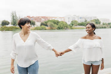 Smiling lesbian couple holding hands at lakeshore - JRVF01943