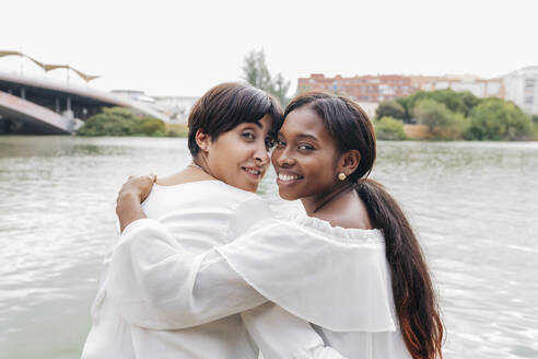 Lächelndes lesbisches Paar schaut über die Schultern auf den See - JRVF01960