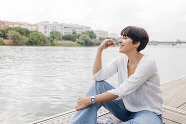 Smiling young woman sitting on pier by lake - JRVF01963