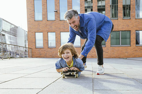 Fröhlicher Vater spielt mit seinem auf dem Skateboard liegenden Sohn - IHF00564
