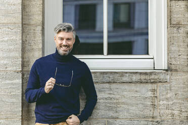 Smiling man holding eyeglasses in front on wall - IHF00584