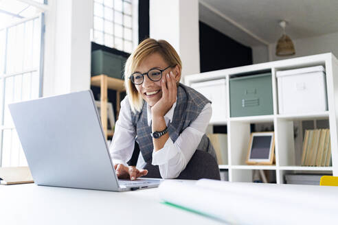Lächelnde Geschäftsfrau mit Hand am Kinn, die einen Laptop benutzt und sich auf den Schreibtisch stützt - GIOF13721