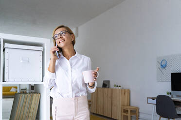 Lächelnde weibliche Fachkraft, die mit einem Mobiltelefon spricht und einen Kaffeebecher im Büro hält - GIOF13735