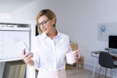 Lächelnde Geschäftsfrau, die eine Kaffeetasse in der Hand hält, während sie mit ihrem Mobiltelefon im Büro Textnachrichten verschickt - GIOF13736