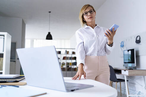 Mittlere erwachsene Geschäftsfrau mit Smartphone am Schreibtisch stehend - GIOF13739