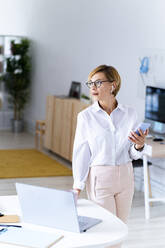 Female professional holding mobile phone while standing at desk - GIOF13740