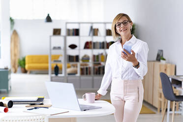 Smiling female professional wearing eyeglasses while holding smart phone at desk - GIOF13741