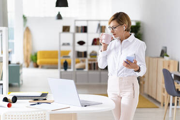 Businesswoman with mobile phone drinking coffee in office - GIOF13742