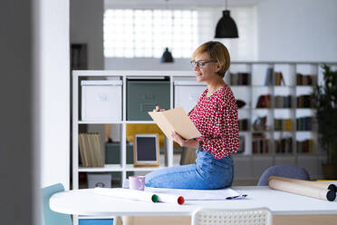 Mid adult female professional writing in diary while sitting on desk - GIOF13748