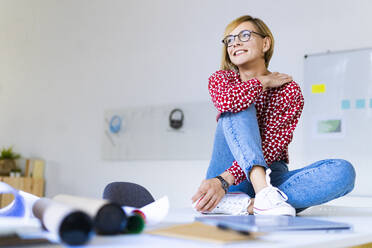 Lächelnde blonde weibliche Fachkraft sitzt auf dem Schreibtisch im Büro - GIOF13764