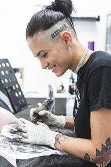Lächelnde Künstlerin beim Tätowieren auf dem Arm eines männlichen Kunden im Studio - JAQF00846